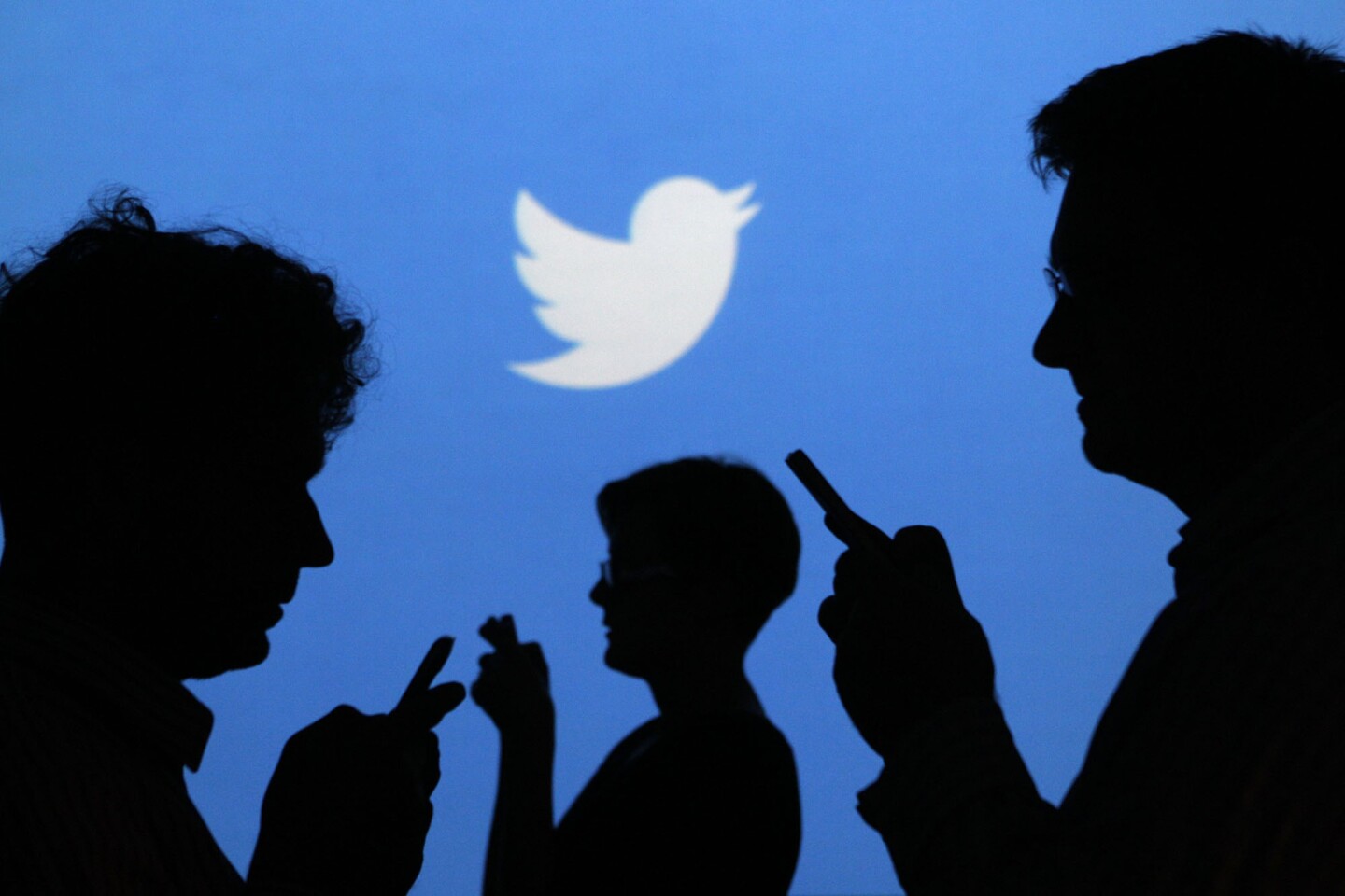People holding mobile phones are silhouetted against a backdrop projected with the Twitter logo in Warsaw